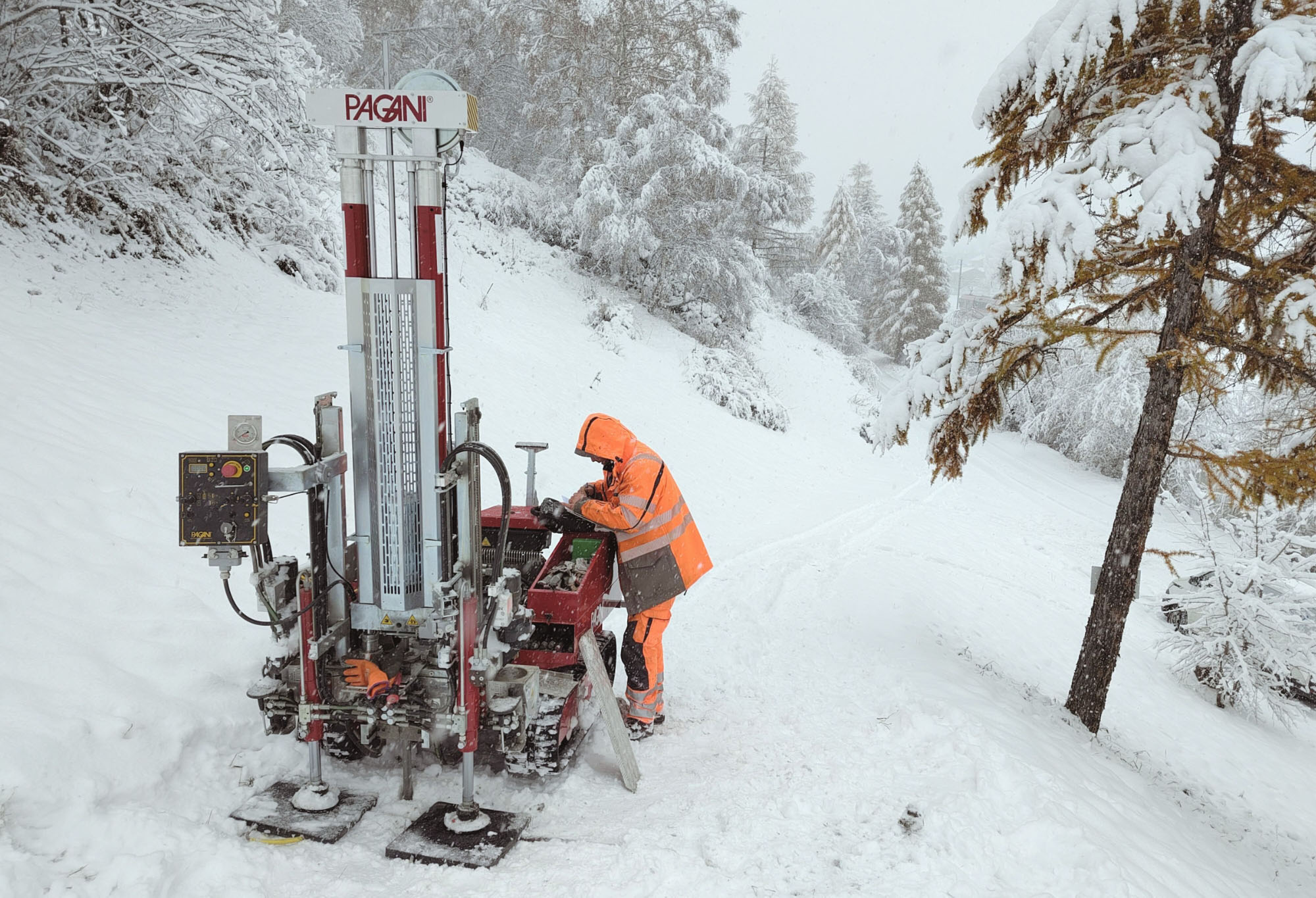 Bohrgerät im winterlichen Gelände: Eine Person in orangefarener Schutzkleidung bedient eine Pagani-Bohranlage auf einer schneebedeckten Strasse, umgeben von stark verschneiten Bäumen und Hangflächen.
